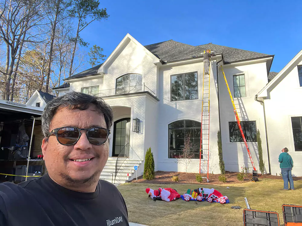 Smiling technician in sunglasses taking a selfie in front of a white house with ladders set up for holiday light installation, with inflatable decorations on the lawn.