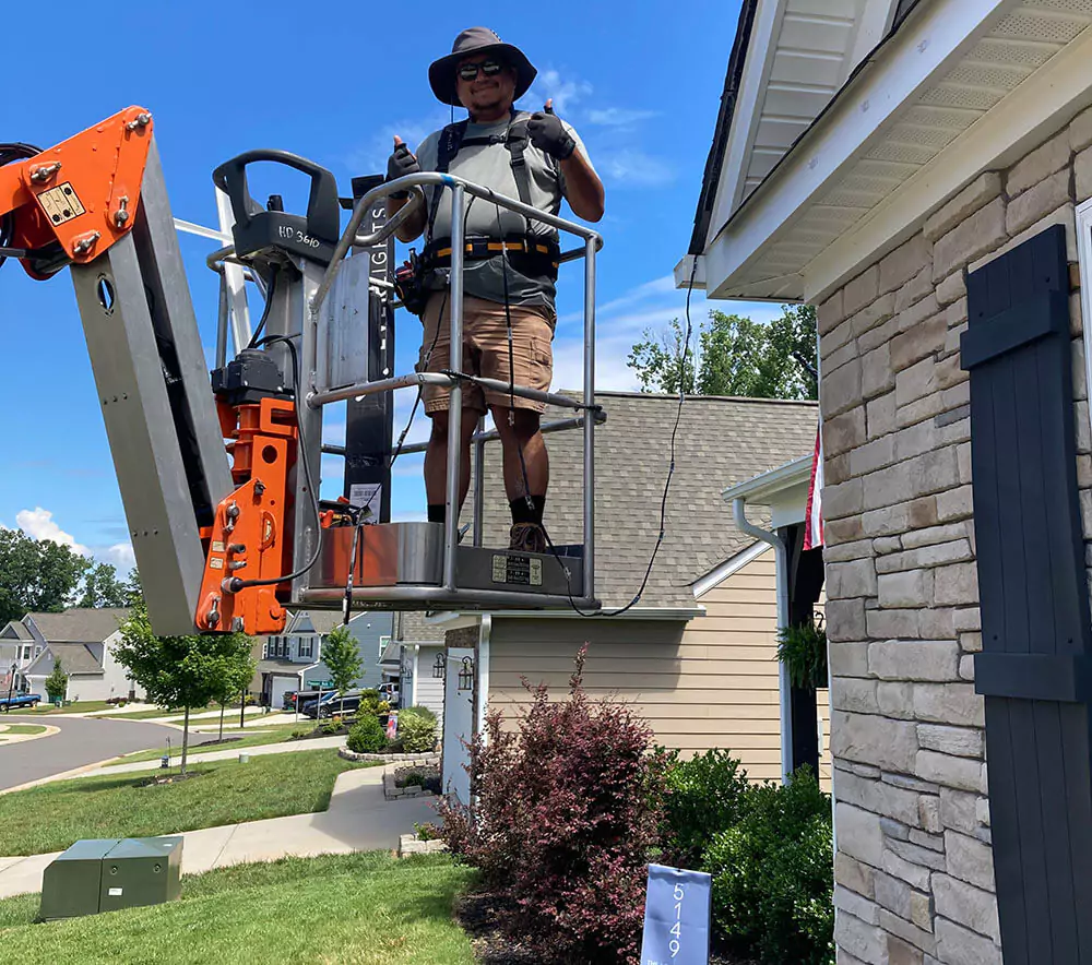 Technician in a lift giving thumbs up while working on exterior home maintenance, wearing safety gear and a sun hat on a bright, sunny day.