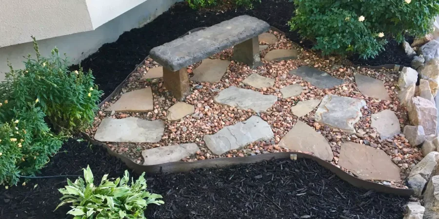 Stone sitting area with rustic bench, flagstone pavers, decorative gravel, and surrounding mulch in a landscaped corner