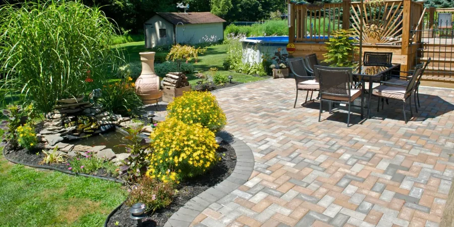 Backyard patio with paver flooring, outdoor dining set, water feature with stone pond, vibrant flower beds, and above-ground pool with wooden deck