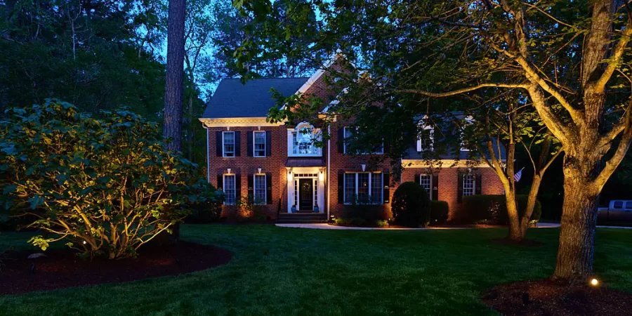 Elegant two-story brick house with landscape lighting highlighting the front yard trees, shrubs, and the house facade during twilight in a wooded neighborhood.