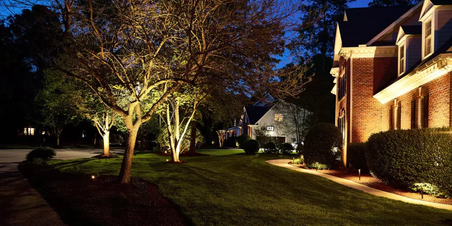 Front yard view of a brick house with landscape lighting highlighting trees, shrubs, and the lawn at night along a quiet neighborhood street.