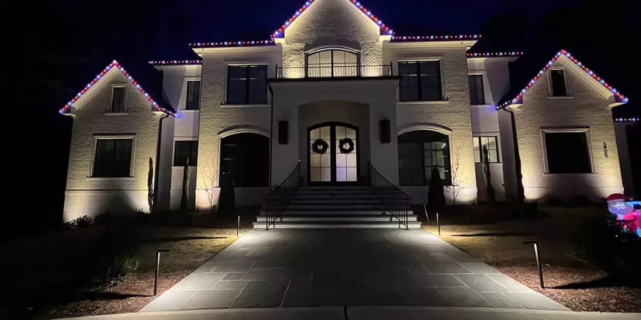 Large white brick house with red and blue roofline lights and festive wreaths on the front doors, illuminated with soft landscape lighting at night.