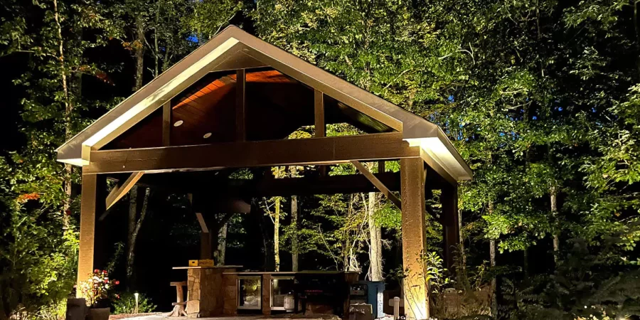 Stylish backyard pavilion with outdoor kitchen, surrounded by lush trees and ambient lighting, creating a serene evening retreat by the poolside.